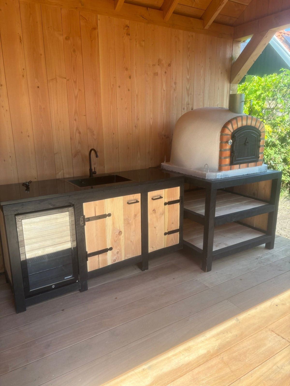 Luxe houten buitenkeuken onder overkapping met ingebouwde witte steenoven, spoelbak en wijnkoelkast