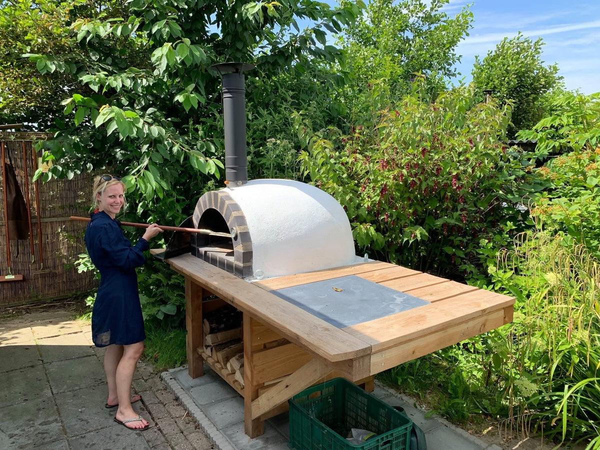 Vrouw bakt pizza met een pizzaschep in een witte steenoven op een houten buitenkeuken in de tuin